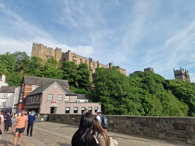This image captures a scene in Durham, United Kingdom, specifically featuring Durham Castle and Durham Cathedral atop a forested hill. A bridge made of stone is in the foreground, with pedestrians walking across. On the left, buildings line the street, one of which has a sign that reads "Akarsu." People are walking in both directions on the bridge. The sky is blue with scattered clouds.