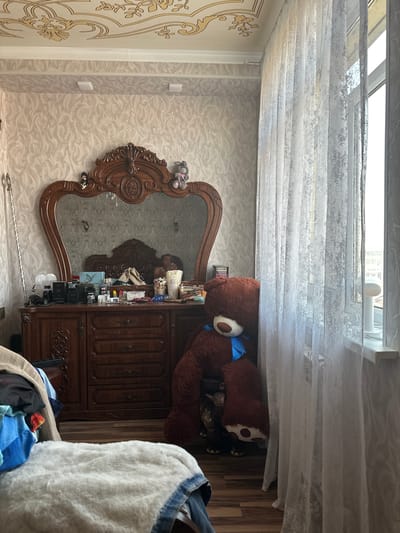 The image depicts an interior scene, possibly a bedroom or dressing area. A large, dark wood dresser with a heart-shaped mirror dominates the center of the frame. The dresser's surface is cluttered with various items including bottles, containers, and what appear to be cosmetics. A very large, plush brown teddy bear with a blue bow is seated to the right of the dresser, partially obscured by sheer white curtains that cover a window. In the foreground, to the left, a pile of folded clothing is visible. The walls are covered in textured wallpaper, and the ceiling features ornate decorative moldings. The floor appears to be wood. The overall impression is one of a lived-in space. No specific details place this scene in Navoiy City, Uzbekistan.