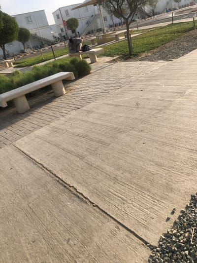 The image depicts an outdoor area in النعيلة, Saudi Arabia, characterized by a mix of hardscaping and sparse landscaping under bright daylight. In the foreground, a textured concrete walkway dominates the lower right, bordered by a strip of gray gravel. To the left, a section of a paved area with rectangular interlocking bricks runs parallel, separated by a light-colored concrete bench. A second identical concrete bench is visible further into the midground.
The midground features a landscaped strip with green grass and low, bushy vegetation. One individual, appearing to be male and dressed in light-colored traditional attire, is seated on the second concrete bench. This person is bent forward, with their head lowered, seemingly attending to a dark bag or backpack placed on the bench in front of them. A slender tree with sparse foliage stands to the right of this person.
The background consists of multiple white, modular buildings, possibly temporary or portable structures, each equipped with windows and external staircases leading to upper levels. Air conditioning units are visible on the sides of some buildings. A red circular sign is partially visible near one of the buildings. A light-colored fabric shade canopy is also partially visible in the upper background, extending over a paved area that appears to be a road or parking lot.