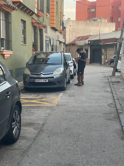 A medium shot captures an alleyway in La Línea de la Concepción, Spain, with a person standing next to an electric scooter and looking at their phone. The scene depicts a narrow street lined with buildings of varying architectural styles. On the left, a building with a green facade features windows with black security bars and terracotta-colored decorative elements. Opposite this, a more weathered building shows signs of age, with a corrugated metal roof.

A dark gray Citroën Picasso car is parked on the right side of the frame, its license plate clearly visible as "9977 FMF". A yellow criss-cross marking on the asphalt indicates a no-parking zone. The person, dressed in black shorts and a black t-shirt, is positioned between the parked car and a white electric scooter. Their posture suggests they are engaged with their mobile device. The ground is paved with asphalt, with a curb and some debris visible on the right side. The overall atmosphere is that of a typical urban street scene in a Mediterranean town.