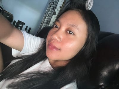 A young woman with black hair and dark eyes is taking a selfie. She is wearing a white t-shirt and has a hair roller in her hair. She appears to be indoors, possibly in a bedroom or living room, as suggested by the presence of a curtain and furniture in the background. The lighting is dim, indicating it might be evening or nighttime. There are no other people visible in the image. The scene is a personal, candid moment captured by the woman herself.