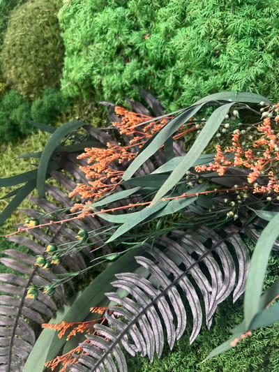 This image displays a close-up of a decorative arrangement of faux greenery and flowers, likely a floral installation or a part of a larger botanical display. The composition features a vibrant, textured green moss as the background, interspersed with various artificial plants. Prominently featured are long, slender green leaves, resembling eucalyptus, and dark, gray-green fern fronds with finely dissected leaflets. Delicate sprays of small, orange-red flowers and buds add a contrasting pop of color and texture. Some green buds, possibly from a different floral element, are also visible. The lighting is even, suggesting an indoor setting or diffused daylight. There is no discernible text within the image, and no people or specific activities are depicted. The overall impression is one of lush, naturalistic, but artificial, botanical beauty. The context of "Minsk, Russia" doesn't provide specific visual cues within the image itself, as the arrangement is a common decorative element.