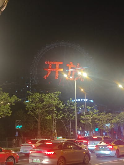 This image depicts a night scene in Nanhai District, China, likely on a city street. In the background, a large Ferris wheel dominates the sky, illuminated with red neon lights forming Chinese characters. To the left of the Ferris wheel, a tall building with visible windows is present. The foreground shows a traffic intersection with several cars waiting. The cars are modern sedans and SUVs, with their taillights illuminated. Traffic lights are visible, indicating a functioning intersection. Trees line the street, adding greenery to the urban environment. Streetlights are also present, contributing to the overall illumination of the scene. The sky is dark, consistent with a nighttime setting.