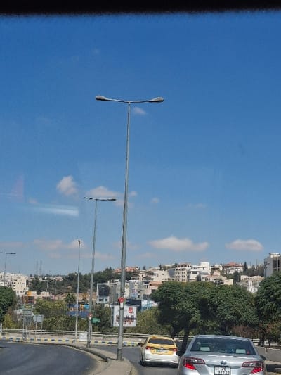 The image captures a daytime urban street scene in Amman, Jordan, from the perspective of a vehicle. The foreground features a curving asphalt road with a concrete barrier displaying yellow and blue markings. Two vehicles are visible: a silver sedan in the immediate right foreground with a Jordanian license plate "20 97727," and a yellow and white taxi sedan positioned slightly ahead. Multiple tall, grey metallic streetlights with double horizontal arms line the road's median. Behind these street fixtures, green trees and foliage are present. The background shows a dense cityscape of numerous light-colored buildings, characteristic of Amman's architecture, situated on rolling hills. The upper half of the frame is dominated by a clear blue sky with several scattered white cumulus clouds. No individuals are depicted.