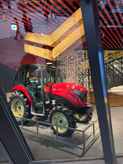 A red Yanmar tractor is displayed indoors, possibly in a showroom or exhibition space. The tractor is positioned on a metal stand. The environment appears modern, with reflective surfaces and architectural elements. In the background, through a glass partition, a reflection shows people walking on a walkway with red striped railings overhead, suggesting an urban setting. There is also a decorative display of what appears to be small, falling pink shapes, possibly representing cherry blossoms. The overall impression is of a product display within a contemporary urban environment. This scene is located in Chuo, Japan.