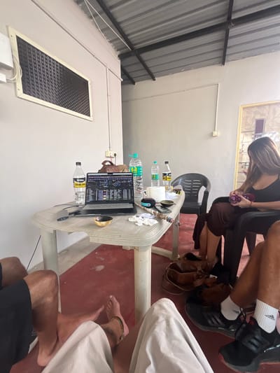 The image depicts an indoor scene in Sanapur, India, featuring multiple individuals gathered around a light-colored plastic table. The room has light grey painted walls, a corrugated metal roof with visible black support beams, and a red-painted concrete floor. Electrical conduits run along the walls and ceiling. A rectangular vent or window frame with a dark mesh is affixed to the left wall. A doorway on the right shows a decorative frame and a patterned inner surface.

On the table, a black laptop is open, displaying what appears to be DJ software. Connected to the laptop is a silver external device. Several water bottles are present, including Kinley Strong Soda and Bisleri branded bottles. A brown leather bag, a small wooden bowl, a roll of white paper, a portable speaker, an ashtray, and a string of beads are also arranged on the table.

At least four individuals are partially visible. In the bottom-left, a person wearing black shorts has bare feet, one adorned with an anklet. Another person in the bottom-center wears light-colored pants and is also barefoot. Seated in a black plastic armchair on the right is a female with long brown hair, wearing a black top and a dark brown skirt. She holds a small, colorful object in her hands. In the bottom-right, a person wears black shorts, black sneakers with white soles, and white socks. A brown leather bag rests on the floor beneath the table.