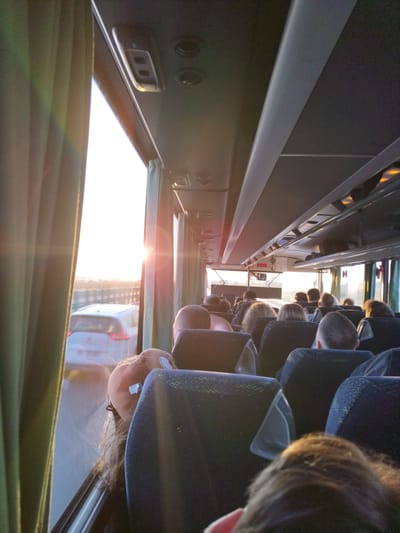 The image is taken from inside a bus, looking forward and slightly to the left. The sun is setting, casting a warm glow through the windows and creating lens flares. On the left side of the image, green curtains are partially drawn. Through the window, a white car is visible on a road, with other vehicles and a distant landscape in the background. The interior of the bus is populated with passengers seated in rows of dark blue seats. The focus is on the aisle and the backs of the seats, with the heads and shoulders of several passengers visible. In the foreground, a plush toy is resting on a seat. The overall atmosphere is one of travel during sunset.
