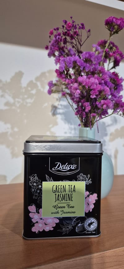 A close-up, eye-level shot showcases a black metal tin of "Deluxe Green Tea Jasmine" tea, positioned on a wooden surface. The tin is adorned with delicate line drawings of pink flowers and green tea leaves, with a light green label displaying the product name in black lettering. A small circular seal with a frog and the words "Rainforest Alliance People & Nature" is visible on the lower right side of the tin. Behind the tin, a vase containing a bouquet of small, vibrant purple flowers is partially visible. The background consists of a textured, light-colored wall with a soft shadow pattern cast upon it, likely from the flowers and an unseen light source. The overall scene suggests a still-life composition, possibly within a home environment. The image does not provide any specific information about its location in Póvoa de Varzim, Portugal.
