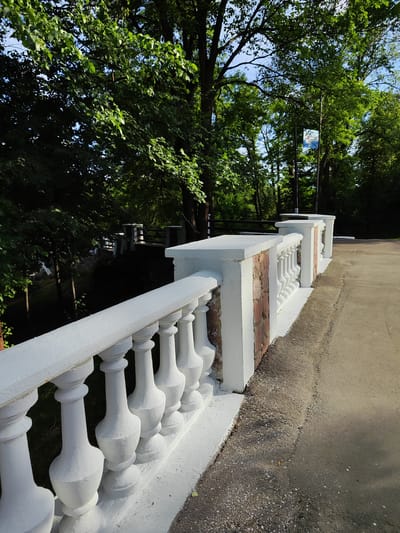 The image depicts a sunny day in a park-like setting in Korosten, Ukraine. The foreground is dominated by a white balustrade with ornate pillars, suggesting a bridge or elevated walkway. The balustrade is made of concrete and shows some signs of age, with a few chipped paint areas. The pillars are spaced evenly and have a decorative, bulbous shape.

Beyond the balustrade, lush green trees fill the background, their leaves catching the sunlight. The foliage creates a dappled effect, with patches of shadow and bright light. In the distance, other sections of balustrade and what appears to be a wooden fence can be seen, also surrounded by trees. A signpost with a reflective surface is visible among the trees, its content unclear due to the distance and angle.

The ground next to the balustrade is a mix of asphalt and gravel, indicating a pathway or road. The overall impression is of a serene and natural environment, likely a public space enjoyed for recreation. The bright sunlight and the vibrant green of the trees suggest it is daytime, possibly late morning or afternoon. There is no visible text of significance. The scene evokes a sense of calm and tranquility.