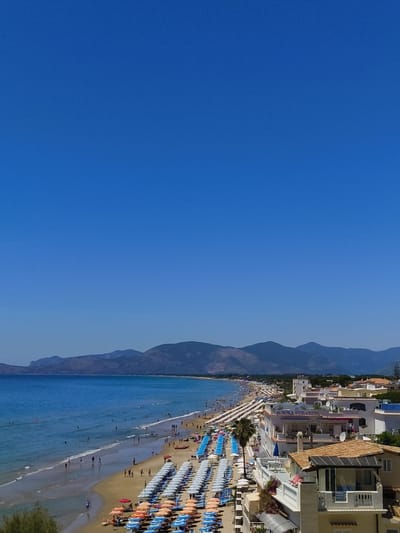 The image shows a vibrant beach scene in Sperlonga, Italy, under a clear blue sky. The foreground features rows of colorful beach umbrellas and lounge chairs, some set up on the sandy shore, others on terraces of buildings. A palm tree stands tall amidst the umbrellas. Beyond the beach, the turquoise Mediterranean Sea stretches out, with numerous people swimming, wading, and enjoying the water. The beach itself is populated with many individuals walking, relaxing, and sunbathing. In the background, a long, undulating coastline is visible, with buildings nestled along the shore and mountains rising majestically behind them. The "AURORA HOTEL & SUITES" is prominently displayed on one of the buildings. The scene captures a lively summer day in a popular coastal resort town.