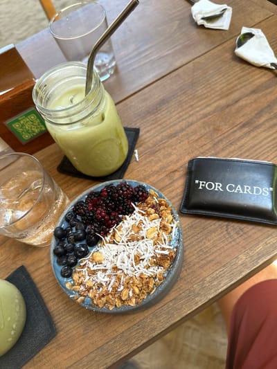 On a wooden table, potentially in a cafe or restaurant in Puerto Escondido, Mexico, sits a colorful array of food items. From top left to bottom left, there are two pieces of silverware wrapped in white napkins, a small black card holder with the words "FOR CARDS" printed on it, and a smoothie bowl with granola, coconut flakes, blackberries, and blueberries. Next to the smoothie bowl is a green drink in a glass jar with a metal straw, an empty glass, and a small green dish on a gray napkin. A person's leg is visible in the upper right corner of the image. The overall scene suggests a meal or snack being enjoyed in a casual setting.