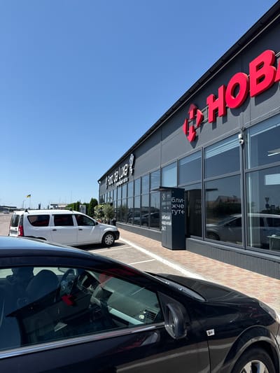 The image shows a modern commercial building with large windows and a dark gray facade, located in Sarny, Ukraine. A large red sign with Cyrillic letters, "НОВ" (NOVA), is prominently displayed on the building's upper section. To the left of the red sign, a logo resembling a stylized arrow or a heart is visible.
In front of the building, a paved area with red and beige bricks lines the entrance. A black car is parked in the foreground, angled to the right, showing its interior with a steering wheel and dashboard. To the left of the black car, a white van is parked, facing left.
A dark gray kiosk or parcel locker is situated near the building's entrance, featuring Cyrillic text that reads, "БЛИЖЧЕ ТУТ" (CLOSER HERE) and "ЗАБИРАЙ НАДСИЛАЙ 24/7" (PICK UP SEND 24/7). The building also has a sign that says "Flora de Luxe" with a flower logo, and below it, "супермаркет квітів" (flower supermarket) in Cyrillic.
The sky is clear and blue, indicating a sunny day. In the background, there are some trees and other buildings, as well as what appears to be a road and possibly infrastructure. A Ukrainian flag on a pole is visible in the distance to the left. Security cameras are mounted on the building.