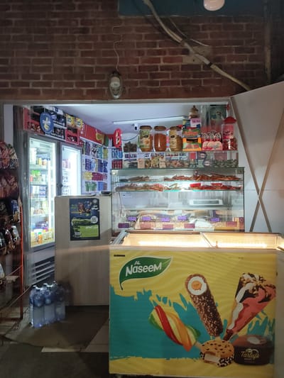 The image shows a small convenience store, likely a "mini-market," in Ariana, Tunisia. The store has a brick facade and appears to be open for business, with its interior lit and stocked with various goods.
Dominating the foreground is a large, bright yellow and teal freezer with colorful ice cream illustrations on its front. The text "AL Naseem" is prominently displayed in green above the illustrations, suggesting it's a brand of ice cream.
To the left of the freezer, a glass-doored refrigerator is visible, filled with bottled drinks and other chilled products. Several large bottles of water are stacked on the floor in front of this refrigerator.
Behind the freezer and refrigerator, the interior of the store is visible, featuring shelves stocked with packaged snacks, candies, and other convenience items. A glass display case is set up, showcasing more items, possibly pastries or other food products. Some of these items have price tags visible.
The overall impression is a typical street-side convenience store in a Tunisian city, offering a range of chilled goods, snacks, and potentially some prepared foods. The lighting suggests it might be evening or nighttime.