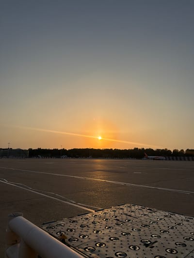 This is a vertically oriented, close-up shot of an airport tarmac during sunset. The sun, a bright orb casting a warm golden glow, is positioned low on the horizon, creating a lens flare that streaks across the image. The sky transitions from a deep blue at the top to a soft orange and yellow near the sun. The tarmac, wet and reflecting the sunset, is marked with white lines, indicating parking or taxiing areas.
In the mid-ground, a line of trees forms a dark silhouette against the sky. Partially visible amongst the trees is the tail of an orange and white airplane, with the "easyJet" logo clearly discernible. The airplane suggests the setting is an operational airport. Red and white striped barriers line the edge of the tarmac near the trees.
In the foreground, on the right side of the image, a large, dark, metallic object with a grid of circular indentations and visible mounting hardware dominates the frame. This appears to be some form of industrial equipment or platform, possibly related to ground support at the airport. A white pipe extends from the bottom right corner of the frame, partially obscuring the metallic object.
The overall impression is one of quiet activity at an airport as the day winds down. The wet tarmac and the angle of the sun suggest recent rain or a damp atmosphere. The time of day is clearly sunset, indicated by the sun's position and the warm lighting. The location is specified as Crawley, United Kingdom. No people are visible in the image.