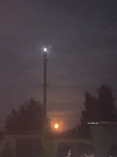 The image depicts an outdoor scene at night, possibly in Zviahel, Ukraine. The sky is dark and appears overcast, with diffused light suggesting a cloudy or foggy condition. A bright, artificial light source is visible in the upper left portion of the frame, possibly a street lamp or the beam of a vehicle. In the middle ground, a tall, slender structure, likely a utility pole or transmission tower, stretches across the image.
Further to the right, a distinct, warm-toned light emanates from what appears to be another street lamp, illuminating the surrounding darkness with an orange glow. Silhouettes of trees with dark foliage are visible against the dimly lit sky, adding depth and context to the scene. The overall impression is one of a quiet, possibly urban or semi-rural setting at night, with artificial lighting providing the primary visual points of interest. The image is blurry, with the focus not sharp, and the colors are muted due to the low light conditions. No people are visible in the frame, and there is no discernible text.