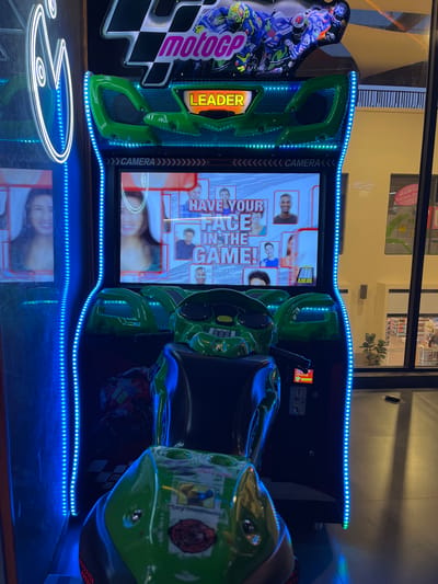 The image shows a "MotoGP" arcade game machine with a motorcycle seat, positioned indoors. The game screen displays a feature to "HAVE YOUR FACE IN THE GAME," showcasing images of people's faces. The machine is illuminated with bright blue LED lights along its edges and base. The arcade is located in Mechouar Kasbah, Morocco. The image features a bright white neon light design on the floor, which is reflecting on the floor's surface. There are various stickers and decorations on the arcade game. A glimpse of a storefront can be seen in the background, through the exterior windows.