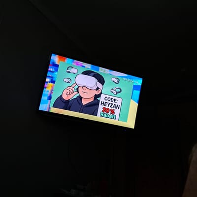 The image shows a close-up of a digital screen displaying an advertisement. The ad features a cartoon character wearing a VR headset, with several smaller cartoon VR headsets scattered around the character. A sign held by the character displays the text "CODE: HEYZAN 20% Rabatt" (Rabatt means discount in German). Above the character, the word "WERBUNG" (advertisement in German) is visible. The background of the advertisement consists of a blue and yellow color scheme with some abstract shapes. The screen itself is placed on a dark surface, and the surrounding environment is dimly lit, suggesting the photo was taken in a low-light setting, possibly indoors. The screen is angled slightly, with some digital artifacts or glitches visible along the edges, particularly on the left side. The overall impression is of a promotional graphic for a product or service related to virtual reality, likely targeting a German-speaking audience.