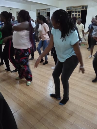 This media file captures a group of people, primarily adults, engaged in an energetic indoor activity that appears to be dancing or group exercise. The scene is set in a spacious room, possibly a community hall or a large studio, featuring a light-toned wooden floor with tile-like divisions and pale walls. Several windows with white frames are visible in the background, allowing for natural light, though artificial lighting may also be in use.
In the foreground on the right, a woman with dreadlocks, wearing a light blue t-shirt, dark leggings, and black and blue socks, is actively moving with her body turned to the left. In the mid-ground on the left, another woman with long dark hair, dressed in a light pink top, colorful patterned pants, and light green pointed flats, is also in motion with her arms crossed. Further to her left, a woman in a black top and black leggings with a red stripe is visible.
Numerous other individuals, both men and women, are participating in the background, some appearing blurred due to movement, indicating a dynamic group activity. Most participants are dressed in casual attire such as t-shirts, jeans, and leggings. Some are seen wearing socks directly on the wooden floor, while others have shoes. The atmosphere suggests a lively and communal event. There is no visible text in the image. The location context indicates this event is taking place in Riara Ridge, Kenya.