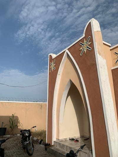 A low-angle shot captures the side of a building, featuring a textured brown facade with decorative cream-colored trim. Two intricate star-shaped embellishments, one in the foreground and another partially visible above, adorn the brown surface. The building's design includes a curved, recessed alcove. To the right, a portion of a tiled floor is visible, along with a black motorcycle and several pairs of flip-flops. A broom leans against the wall near the motorcycle. In the background, dry vegetation grows. A section of barbed wire fencing runs along the top of a beige wall. The sky above is a clear blue, dotted with wispy white clouds. The scene appears to be set in Wai A, Nigeria. The lighting suggests it is daytime, likely late afternoon or early morning, given the shadows. The overall mood is neutral, presenting a snapshot of an outdoor scene with architectural and everyday elements. No visible text is present in the image.