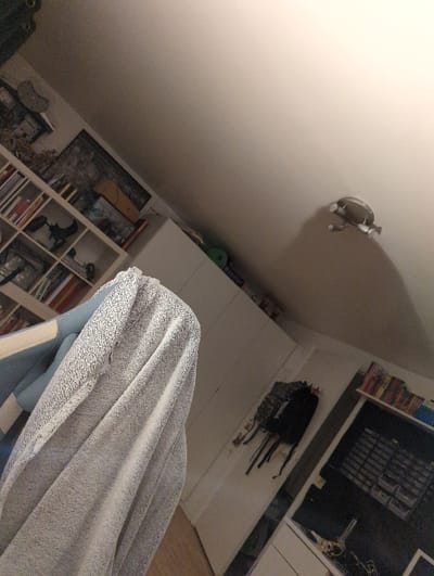 The image shows a white towel, a light gray blanket, and a white door. There are also bookshelves on either side of the door with books, a backpack hanging on hooks, and a shelf with a variety of objects. The ceiling is white with a light fixture in the middle. The location is a bedroom in Lagny-sur-Marne, France, at coordinates 48.879, 2.705. The photo was taken at an upward angle.