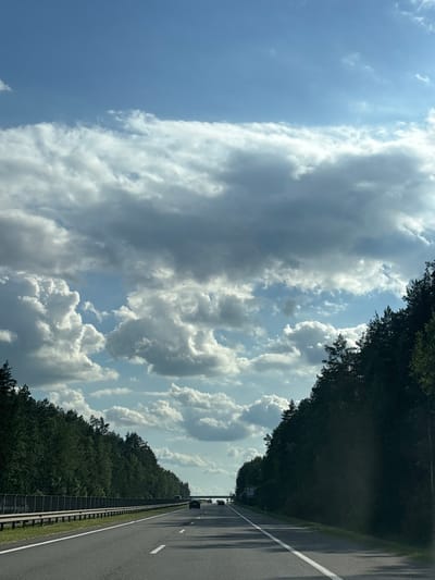 The image shows a road with a dense line of trees on the left and the sky filled with clouds on the right. The road appears to be a highway with a white line marking the lanes. There are a few cars visible on the road in the distance. The trees are tall and lush green, creating a natural barrier along the roadside. The sky is a mix of blue and white, with scattered clouds of various sizes and shapes. Some clouds are dark and ominous, suggesting the possibility of rain, while others are bright and fluffy, illuminated by the sun. The overall scene evokes a sense of travel and nature. The time of day appears to be daytime, likely afternoon, given the lighting conditions. There is a sign with text in a foreign language visible near the road, but it is too small to be legible. The location is specified as Драчкаўскі сельскі Савет, Belarus.