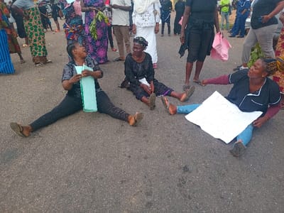 The image depicts a scene in Ungwan Tanko, Nigeria (latitude: 10.422, longitude: 7.49). Four women are sitting on a paved street, engaged in a protest. Two of the women are seated in a traditional sitting position, with their legs crossed. The woman on the left holds a light blue paper in her hands. She is wearing black leggings and a black shirt. The woman on the right is sitting in a similar position and holds a blank piece of paper in her hands. She is wearing blue jeans and a black shirt. The woman in the center, wearing a black and white dress, looks distressed as she holds a green cloth in her hands. The fourth woman is wearing black leggings and a black shirt. She is sitting in a seated position with her legs spread apart, holding a folded light blue paper. Several people are standing in the background, seemingly observing the situation. It appears to be a public space, potentially a road or a street, with a crowd gathered around the women sitting on the ground. The scene conveys a sense of unrest and protest.