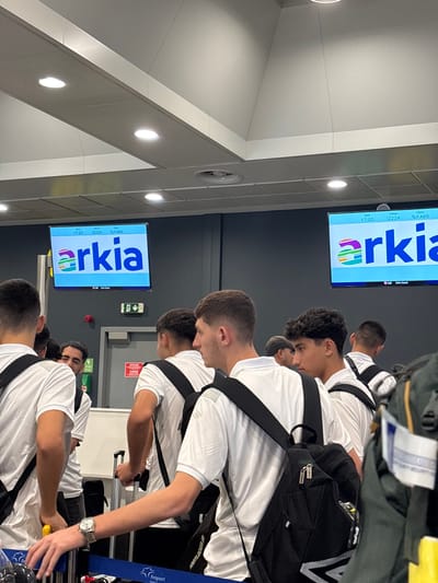 The scene depicts an indoor airport terminal, plausibly Thessaloniki Airport 'Makedonia' (SKG), which serves the region encompassing Νέο Ρύσιο, Greece. Multiple young males are observed standing or moving forward, forming a queue. They wear white short-sleeved polo shirts and predominantly carry black backpacks. One individual in the immediate foreground has a silver-colored wristwatch on his left wrist. Another individual in the mid-ground operates a piece of rolling luggage with a yellow handle.

The background features a dark grey wall with two large, illuminated digital display screens mounted high, each showing the 'arkia' airline logo and name. Textual information on the screens includes '17:20', '12224', and 'Τελ Αβίβ' (Tel Aviv), indicating flight details. Between the screens, a red fire alarm and a green exit sign are present above a grey door. The ceiling is white, featuring multiple recessed circular light fixtures. In the foreground, queue management barriers with blue retractable belts are visible; one belt is marked with 'Fraport Greece'.