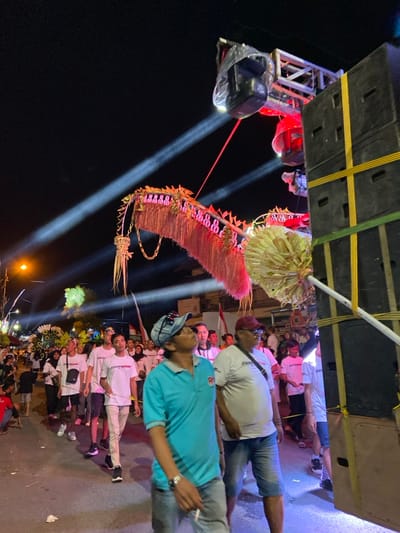 The image depicts a nighttime procession on a street in Kedungbanteng, Indonesia. In the foreground on the right, a tall stack of dark-colored PA speakers with yellow strapping is present, adjacent to a metal truss system supporting multiple moving head stage lights, some emitting powerful white beams into the dark sky, others casting red light. One light fixture is partially covered in clear plastic.

In the immediate foreground, two adult males are walking right-to-left. The man closest to the camera wears a light blue polo shirt, blue jeans, and a light-colored cap, looking upwards and to his right, holding a lit cigarette in his right hand. Beside him, another man wears a white t-shirt, denim shorts, a dark cap, and a shoulder bag, looking forward.

The midground features a dense procession of individuals, predominantly young adults, many wearing white t-shirts. They are walking in the same direction as the foreground figures. Prominently displayed within the procession are large, elaborate decorative structures. One significant object is a long, curved, dragon-like effigy adorned with reddish-pink fringed material and green accents, likely constructed from woven natural fibers or fabric. Another decorative object, resembling a large, fanned whisk, is held upright near the speaker stack. Additional similar decorative elements are visible further down the street.

The background reveals a large gathering of people, some indistinct buildings, and streetlights. Faint red and white flags, consistent with the Indonesian national flag, are visible among the crowd. The ground near the speaker stack is illuminated with a purple-blue light. The overall scene is brightly lit by artificial event lighting and streetlights against a dark sky.