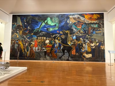 A large, multi-panel mural dominates the central view of an indoor exhibition space in San Juan, United States. The mural, rendered in a dynamic, social realist style, depicts a complex, allegorical scene with numerous human figures, a large green spectral being reaching downwards, a dark rearing horse in the center, skeletal figures, industrial machinery, and a prominent red, horned face with flames in the upper right. Figures in the foreground of the mural appear to be in crowds, some interacting, others in distress or struggle, while structures behind them are shown in various states of collapse or construction.
The exhibition space features a polished light-brown hardwood floor that reflects the overhead lighting. The walls are plain white. In the lower left foreground, a section of a white pedestal displaying a silver, abstract, curvilinear sculpture is visible. Several silver-colored stanchions, some with attached information placards, are positioned along the base of the mural, acting as a barrier. A person, viewed from the back and left side, wearing a dark cap and a green shirt, stands on the far left, facing the mural. A second person is partially visible on the far right.