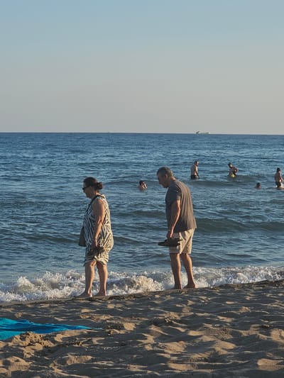 The image depicts a serene beach scene in Castelldefels, Spain, bathed in the warm glow of late afternoon sunlight. The foreground is dominated by a sandy beach, with a vibrant blue towel lying on the sand. In the mid-ground, the gentle waves of the Mediterranean Sea lap at the shore, creating a soft foam. Several people are enjoying the water, with some wading and others swimming. In the background, the vast expanse of the sea stretches to the horizon, where a cargo ship is visible in the distance, adding a touch of industrial presence to the natural beauty.

Two individuals are walking along the water's edge. A woman in a black and white patterned dress walks to the left, her gaze directed downwards. To her right, a man in a grey t-shirt and beige shorts walks to the right, carrying a pair of dark-colored sandals in his hand. The scene is tranquil, capturing a moment of leisure and relaxation on the coast.
