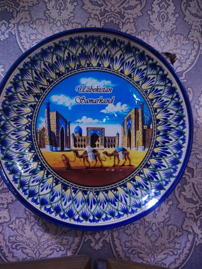 The image displays a decorative plate featuring a scene from Samarkand, Uzbekistan. The plate has a blue rim with intricate white and blue floral patterns. The central design depicts a desert landscape with two camels carrying riders. In the background, traditional Uzbek architecture with blue domes and minarets are visible. The words "Uzbekistan Samarkand" are written in cursive script above the scene. The plate is hung on a wall covered with a patterned wallpaper. The lighting suggests an indoor environment.