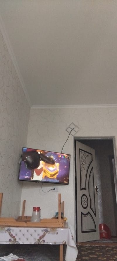 A television is mounted on a wall with wallpaper. The television displays a cartoon with animated characters and a golden box. Below the television, a simple wooden shelf is set up on a table covered with a patterned tablecloth. On the shelf, a pitcher with a red lid is visible. To the right of the television, a partially open door with decorative carvings is present. A red bucket is located on the floor near the doorway. The room appears to be illuminated by natural light, and the floor is carpeted with a patterned design. The scene suggests a domestic setting within a room in Zafarabad, Uzbekistan.