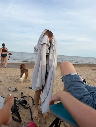 A family is enjoying a day at the beach in Ondvor, Russia. The scene is set on a sandy beach with the ocean in the background. A woman in a black bikini walks towards the water, carrying a paddle. A brown and white dog, likely a Spaniel, stands nearby on the sand, facing away from the camera. In the foreground, a child is wrapped in a grey towel, obscuring their face. To the right, a person's legs are visible, clad in ripped denim jeans, resting on a blue beach chair. Several pairs of sandals and other beach accessories are scattered on the sand around the people. The sky is partly cloudy, with the sea appearing calm.