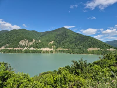 A wide-angle, eye-level shot captures a serene natural landscape in Aranisi, Georgia. The foreground is dominated by lush green foliage, with some wildflowers scattered amongst the grasses. Beyond the greenery, a tranquil, turquoise body of water stretches horizontally across the frame, reflecting the clear blue sky above. The water is bordered by a steep, tree-covered mountain range that recedes into the distance. A road is visible carved into the side of one of the mountains, with a few structures that appear to be some sort of industrial infrastructure located along it. The sky is a vibrant blue, punctuated by scattered white clouds, with the sun casting a bright light on the scene.