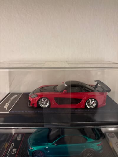The image depicts an indoor scene within the Kajang Municipal Council, Malaysia, showcasing two miniature model cars displayed in clear protective cases.

The primary subject, occupying the upper two-thirds of the frame, is a highly detailed 1:64 scale model of a red sports car, resembling a heavily modified Mazda RX-7. The car features a red body with a matte black hood and roof. It exhibits a widebody kit, aggressive front bumper, prominent side skirts with visible air ducts, a large rear spoiler, and chrome multi-spoke wheels with low-profile tires and noticeable negative camber. This model rests on a dark blue base with white stitching, which includes a rectangular black section on the left displaying a white logo, partially obscured but resembling "MINIGT". The entire upper model and its base are enclosed within a transparent rectangular plastic display case.

Below this, partially visible in the bottom third of the image, is another miniature model car, a teal or dark green four-door sedan, identifiable as an Alfa Romeo Giulia, also within a clear display case. This model features black multi-spoke wheels. A partially visible label on its base reads "Giulia GT".

The background consists of a plain, light-colored (off-white) textured wall directly behind the upper display case. No people are visible. The scene suggests a static collection display.