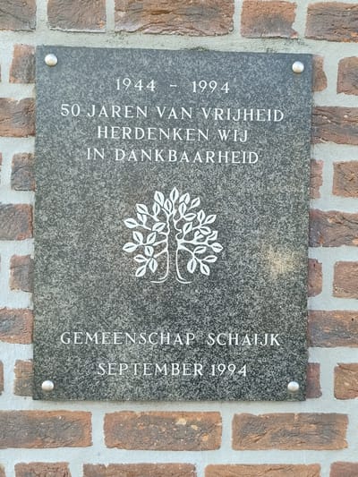 The image shows a memorial plaque mounted on a brick wall. The plaque is made of dark granite with the inscription etched in white lettering. The text on the plaque reads: "1944 - 1994 50 JAREN VAN VRIJHEID HERDENKEN WIJ IN DANKBAARHEID" and below that, "GEMEENSCHAP SCHAIJK SEPTEMBER 1994". This translates to "1944 - 1994 50 YEARS OF FREEDOM WE REMEMBER IN GRATITUDE" and "COMMUNITY SCHAIJK SEPTEMBER 1994".

In the center of the plaque, there is a white stylized illustration of a tree with many leaves, symbolizing life and remembrance. The plaque is secured to the brick wall with four visible metal screws, two at the top and two at the bottom. The brick wall appears to be made of red and brown bricks with light grey mortar. The lighting suggests it is daytime, with no strong shadows visible, indicating possibly overcast weather or indirect sunlight. The overall tone of the memorial is solemn and commemorative, marking a significant historical anniversary. The location is identified as Schaijk, Netherlands.