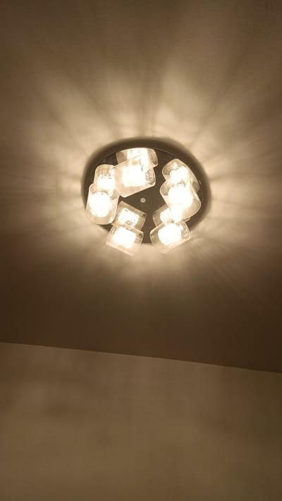 The image shows a close-up, low-angle view of a modern ceiling light fixture. The fixture is circular with a dark base, and it holds multiple clear glass shades, each containing a light bulb. The bulbs are illuminated, casting a warm glow and creating shadows on the textured ceiling. The ceiling itself is a neutral, light color, and it meets a darker, upper wall or ceiling surface at an angle in the upper portion of the image. The lighting creates a somewhat dramatic and atmospheric effect. There are no people, animals, or any other objects visible in the scene. The location in Villaz, France is not discernible from the image itself.