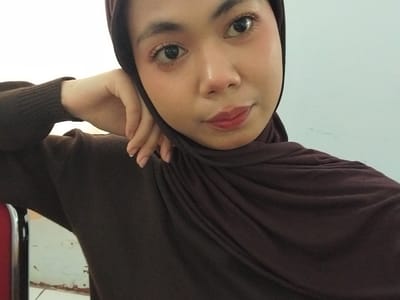 A young woman wearing a brown hijab and sweater leans her head on her hand. Her eyes are open and looking at the camera, and she is wearing makeup. The background is a plain, light blue wall. The image appears to have been taken indoors, possibly in a residential setting. There is no discernible city or location information present in the image.