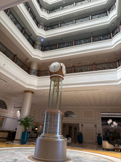 The image shows the interior atrium of a hotel in Ras Al-Khaimah, United Arab Emirates. The atrium features multiple levels with balconies overlooking a central open space. A large, ornate clock tower stands in the center of the atrium, with its glass enclosure revealing internal mechanisms. The architecture is grand and ornate, with white walls, decorative railings, and large columns. Plants in blue planters are placed strategically around the atrium. In the background, there are glimpses of seating areas, a reception desk, and chandeliers, indicating a luxurious hotel lobby. The lighting suggests it is daytime, with light filtering down from the upper levels.
