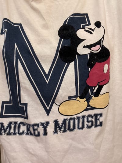 The image displays a close-up of a white t-shirt featuring a graphic of Mickey Mouse and the letter "M". Mickey Mouse is depicted in his classic red shorts and yellow shoes, with a large smile. The letter "M" is in a dark blue collegiate-style font, with white trim. Below the graphic, the words "MICKEY MOUSE" are printed in a similar font. The t-shirt appears to be made of cotton and is slightly creased. There is no discernible location within the city of Novaya Usman', Russia, visible in this image. The photograph focuses solely on the t-shirt's design.