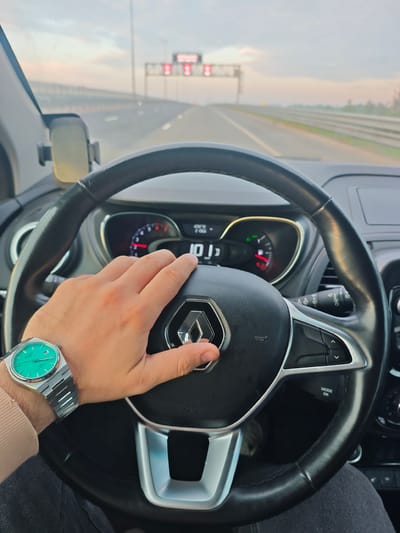 The image shows a first-person perspective from inside a car driving on a highway. The steering wheel, dashboard, and a driver's hand are prominent in the foreground. The hand, wearing a silver watch with a green face, rests on the steering wheel. The dashboard displays digital readouts, including a speed of "101". Outside the car, the highway stretches into the distance, with overhead gantries and blurred trees visible. The sky is overcast with a hint of pinkish light, suggesting either dawn or dusk. The location is identified as Торфяное, Russia.