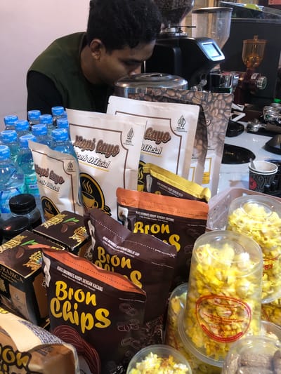 This media file shows a close-up, vertically oriented shot of a person interacting with various products, likely in a retail or market setting. The subject is a person with dark hair and olive-toned skin, wearing a dark green vest over a black shirt. They are positioned on the left side of the frame, leaning in and appearing to be holding or examining a metal object, possibly a coffee cup or container.
The scene is filled with packaged goods. Prominently featured are bags and containers of snacks, including popcorn and what appear to be cookies or brownies, with visible text in Indonesian, such as "Kopi Rayo Aceh Official," "Bron Chips," and "Arabica Gayo." There are also several bottles of water with blue caps lined up at the bottom of the frame. In the background, partially visible, is what looks like a coffee grinder and other coffee-related equipment.
The overall impression is one of a point-of-sale or product display. The lighting suggests an indoor environment, possibly under artificial lights, as there are no strong indications of natural light or weather. The activity seems to be focused on the products, with the person engaged in close observation or handling. The visible text indicates that the products are from Indonesia, with one text snippet mentioning "Tambonbaroh, Indonesia," providing a specific location context. The scene appears to be focused on the sale or promotion of these food and beverage items.
