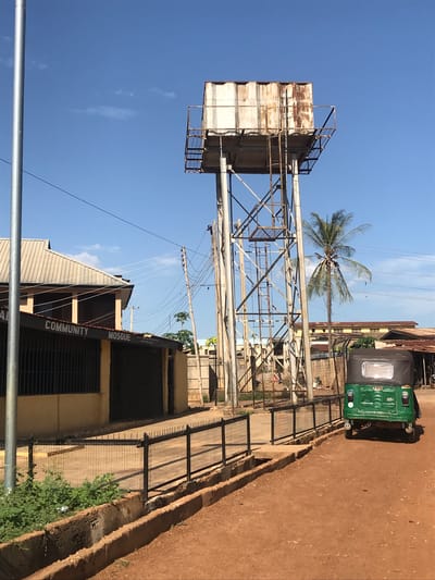 The image presents a sunlit outdoor scene in Mandate Estate, Ilorin, Nigeria, under a clear blue sky dotted with a few faint wispy clouds. The photograph is oriented vertically, showing the ground extending from the bottom-right towards the top.
The dominant feature is a tall, metal-framed water tower situated prominently in the left-center. It supports a large, rectangular water tank that appears weathered, with sections showing discoloration and possibly rust, indicative of age or heavy use. A metal walkway or railing can be seen around the top perimeter of the tank.
Behind the water tower and slightly to its right, a tall, slender palm tree with lush green fronds stands out against the sky. A network of overhead utility poles and power lines stretches across the mid-ground, leading towards a building in the lower-left portion of the frame. This structure features a corrugated metal roof, light-colored walls, and windows protected by dark metal bars. Clearly visible text on the building's facade reads "COMMUNITY" and "MOSQUE," identifying it as a community mosque.
On the right side of the image, a wide, reddish-brown dirt road or unpaved path is present. A green and black auto-rickshaw, commonly known as a 'Keke Napep' in Nigeria, is parked on this road, displaying "TVS" on its front. Metal fences and railings run along the edges of the building and the dirt path, with some green shrubs growing near the base of the fence.
The scene is static, capturing a slice of daily life in a bustling urban or semi-urban environment. The bright illumination suggests it is mid-morning or afternoon.
