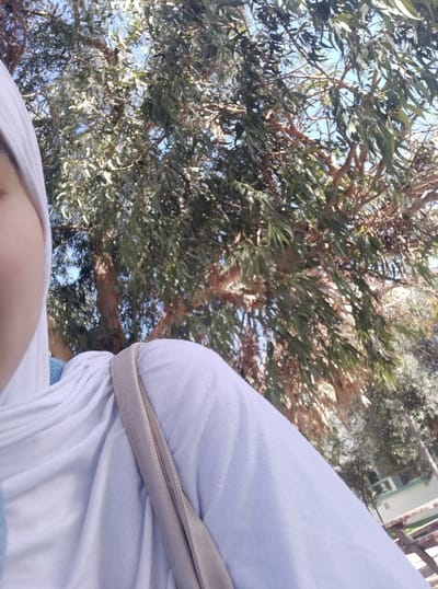 The image shows a person wearing a white top with a gray strap. The view is from below, looking upwards. In the background is a large tree with dense green foliage and brown branches. Patches of bright blue sky are visible through the tree's canopy. The photo is taken during daylight with good lighting.
