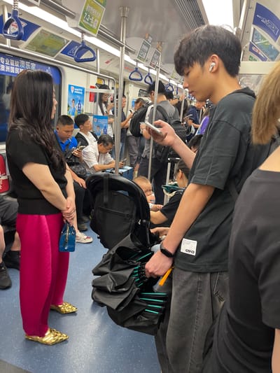 In Longhua District, China, on a subway train, a woman with long dark hair stands on the left, wearing a black top and vibrant pink pleated pants. She holds a small blue handbag in her right hand. Her feet are adorned with gold woven flats. To her right, a young man with short dark hair, wearing a black t-shirt and grey jeans, holds a smartphone in his right hand and has an earbud in his left ear. He is facing away from the camera. A baby in a stroller is partially visible in the foreground. Other passengers are seated and standing throughout the train car, some looking at their phones. The subway car features overhead handrails, advertisements on the walls, and a blue floor.