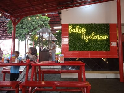 This image depicts a semi-open dining area of a restaurant in Tanjungarum, Indonesia. The foreground features several red wooden tables and matching benches. On the closest visible table, there are multiple condiment bottles (dark liquid, reddish sauce), a white napkin dispenser, a small red ceramic pot, and a promotional stand displaying "500" in white text against a red background.

Seated at a red table in the mid-ground, a woman wearing a patterned dress and a brown hijab looks to her left. Next to her, a young boy in a light blue t-shirt and dark shorts is seated, looking towards his right. Partially visible on the far left, another person's arm and clothing are seen.

On the right side of the image, a prominent wall features a yellow neon sign spelling "Bakso Ngelencer" in script, set against a rectangular panel of green artificial foliage. This panel is framed by red tiles above and below, with a section of black patterned tiles at the base of the wall. Adjacent to the neon sign, a small white sign with red text and a circular stop symbol is affixed.

In the mid-background, behind the seated individuals, a man in a light-colored polo shirt and blue jeans stands near a food preparation or service counter with a dark, sloped roof. Lush green trees are visible in the outdoor background beyond this structure. The overhead structure consists of red metal beams and a white ceiling, from which string lights are suspended. The flooring appears to be dark and textured.