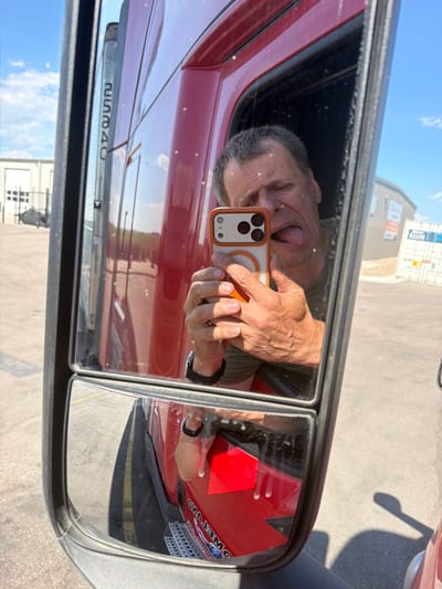 A man is taking a selfie in the side mirror of a red semi-truck. He is sticking his tongue out and making a face. The truck appears to be parked in a lot with other semi-trucks and buildings in the background. The sky is blue with some clouds. The image is taken from a low angle, looking up at the man in the mirror.