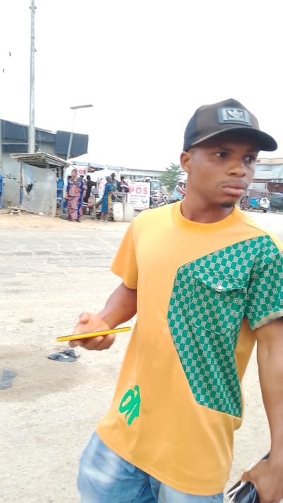 A young man stands on a paved road in what appears to be a busy area in Warri, Nigeria. He wears a brown baseball cap, an orange t-shirt with a green patterned section on the left side, and blue jeans. He holds a yellow object in his left hand and a white object in his right hand.
Behind him, other individuals are visible. There is a small stall or shop on the left, a POS sign can be seen further back, and several vehicles, including what looks like a motorcycle and a three-wheeled auto-rickshaw, are present. The environment seems to be an urban street scene with utility poles and buildings in the background. The weather appears to be overcast.