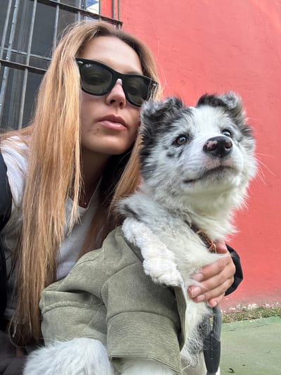 A woman with sunglasses and long blonde hair poses with a fluffy, black and white dog. The dog wears a jacket. The woman's hand rests on the dog. A red wall is behind the dog. The photo appears to be taken outdoors with a metal gate visible. The dog looks up at the camera.