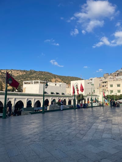 An outdoor, eye-level, wide shot captures a daytime scene in the main square in front of the Mausoleum of Moulay Idriss I in Moulay Idriss Zerhoun, Morocco. The foreground consists of a large, light-colored paved plaza with rectangular stone slabs.
In the mid-ground, a prominent white architectural complex featuring a series of arched walkways with dark interiors and a roofline adorned with green tiles runs horizontally across the frame. Beneath and in front of these arches, striped green and white awnings suggest market stalls or seating areas. Several decorative streetlights with tall, slender green poles and ornate lanterns are spaced along the edge of this complex; some display red Moroccan flags with a green star. Approximately 15-20 individuals are visible in this area, engaged in various activities. One man in a white tunic and dark vest stands facing right, near the center-left. Others are seated on the ground, standing in small groups, or moving through the area, dressed in traditional attire.
The background features a scrub-covered hill rising steeply on the left and center, while a cluster of light-colored, multi-story buildings of the city extends up the hillside to the right. The sky overhead is a clear, deep blue with scattered white cumulus clouds, primarily in the upper right quadrant. Bright sunlight illuminates the scene, casting shadows forward and to the right.
